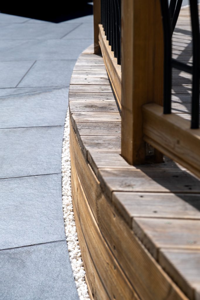 Curved edge of wood decking with white chipping trim next to dark grey tiles. Copyright Molten Marketing