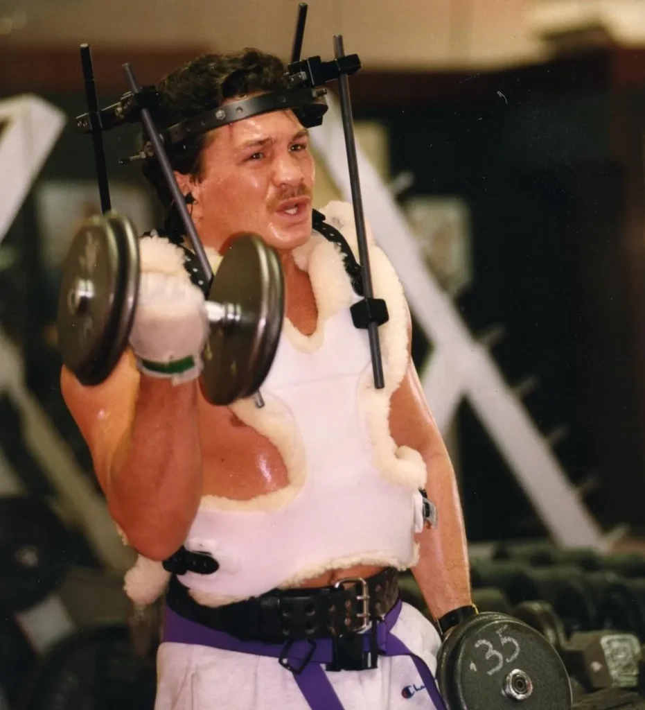 Image of Vinny Paz bicep curling dumbbells with a halo top screwed into his head.
